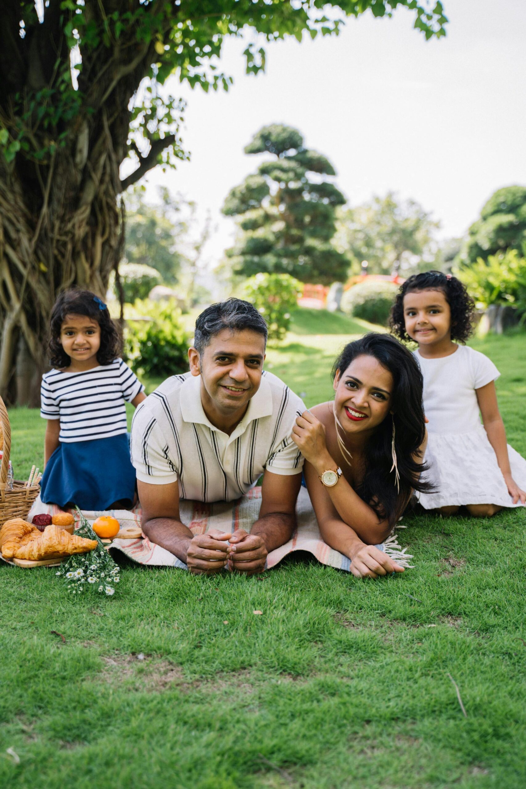Family of four enjoying a picnic in a sunny park with lots of smiles and happiness.